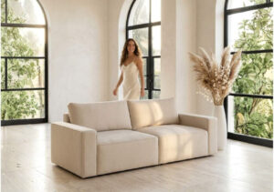 Beżowa sofa modułowa w jasnym wnętrzu z łukowymi oknami, za nią uśmiechnięta kobieta w sukience